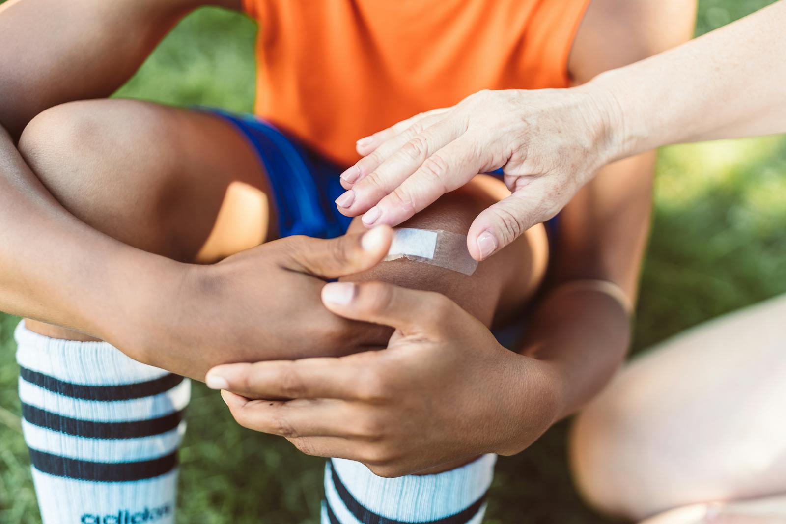 A close-up view of an adult applying a band aid to a child's wounded knee outdoors during the day.